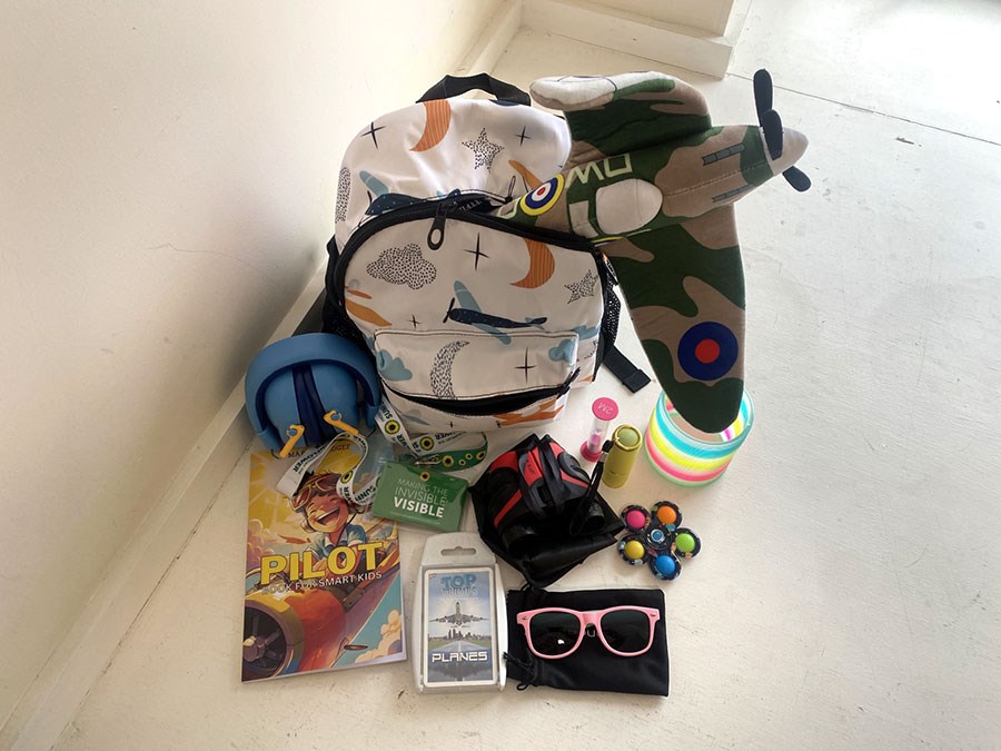 A backpack with several sensory aids surrounding it, including a cuddly toy of a plane, ear defenders, and sunglasses.
