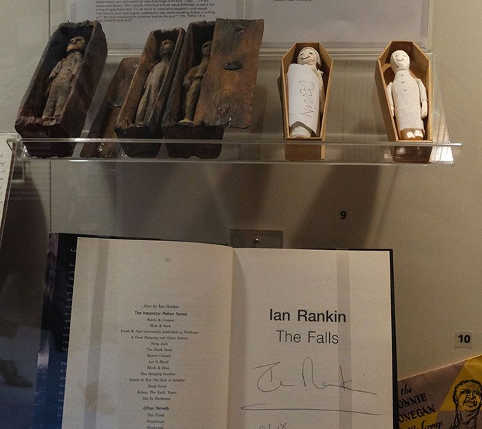 Four Arthur's Seat coffins and two temporary replacements on a plastic shelf above an open book, 'The Fall' by Ian Rankin.