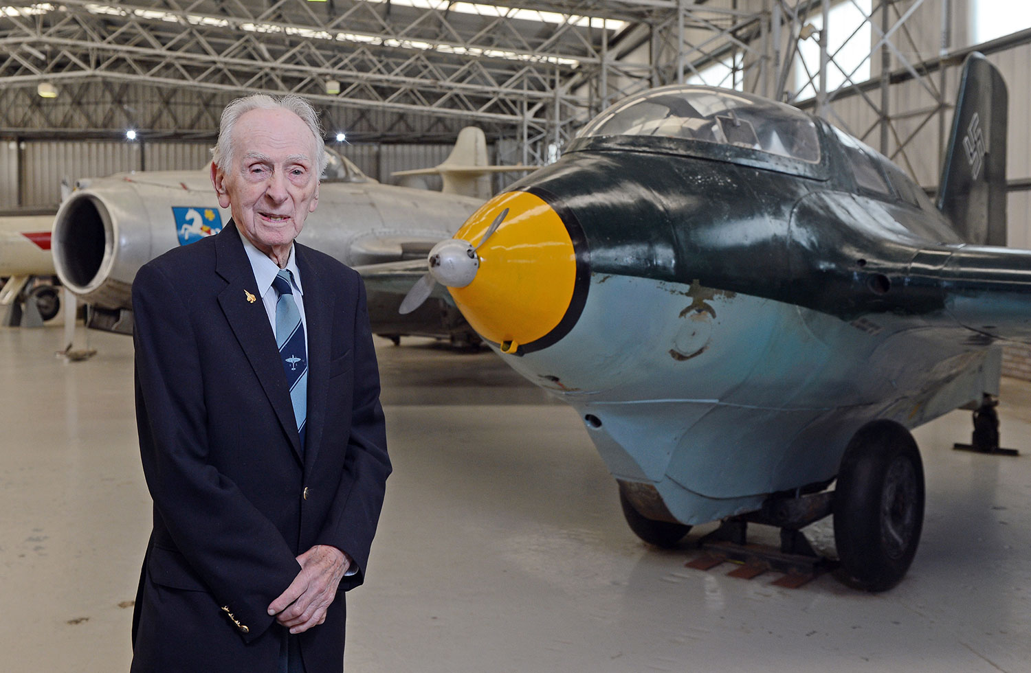 A former pilot, Captain Eric Brown, standing next to a Messerschmitt Komet aircraft in a large aviation hangar.