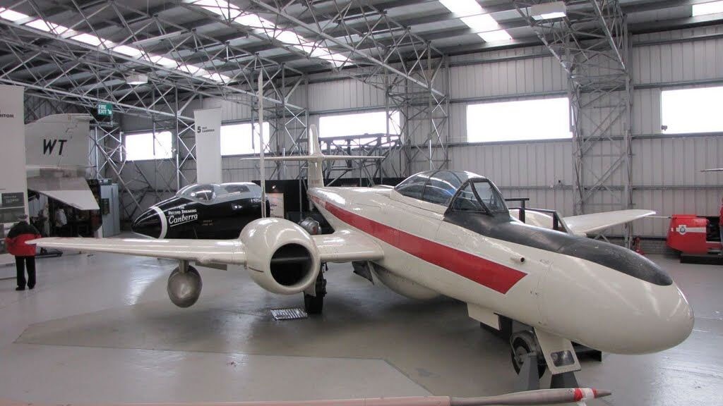 A white Armstrong Whitworth Meteor aircraft with a red stripe parked in a large aviation hangar. There are other aircrafts in the distance.