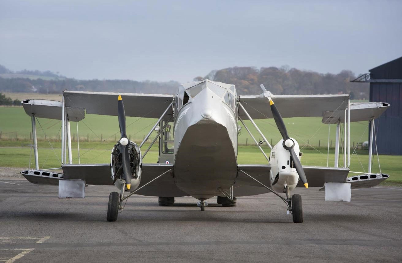 The front side of a grey de Havilland Dragon aircraft on a runway with a hangar and hills in the background