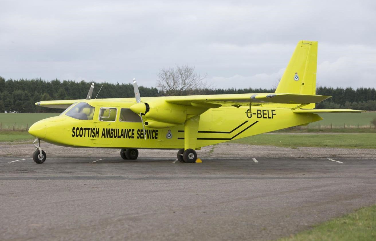 A yellow Britten-Norman Islander aircraft with wide wings parked on a runway. The side of the plane reads 'Scottish Ambulance Service'