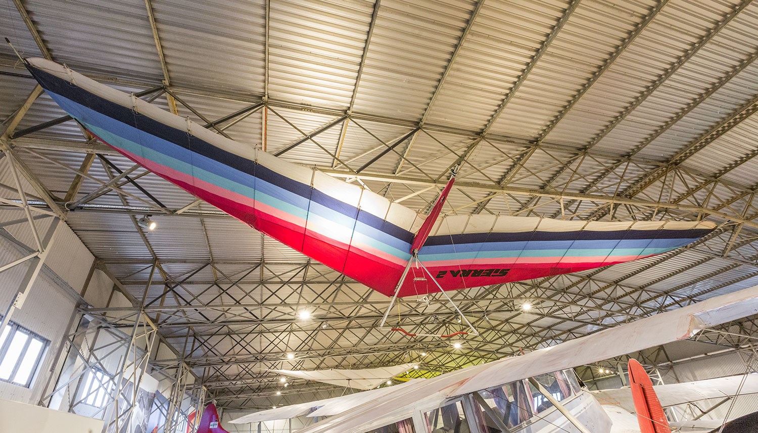 A multi-coloured Firebird Sierra 2 hang-glider suspended fromt he ceiling of a hangar.