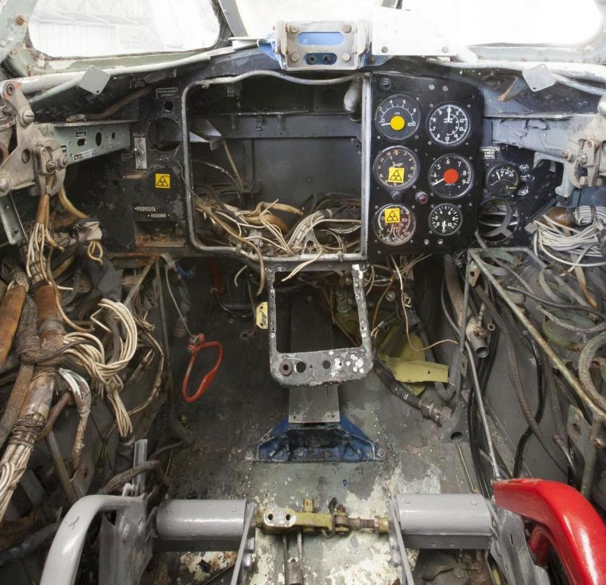 Inside the cockpit of an Aero S-103 aircraft. Parts have been stripped, exposing many wires. There are six dials on the dashboard.