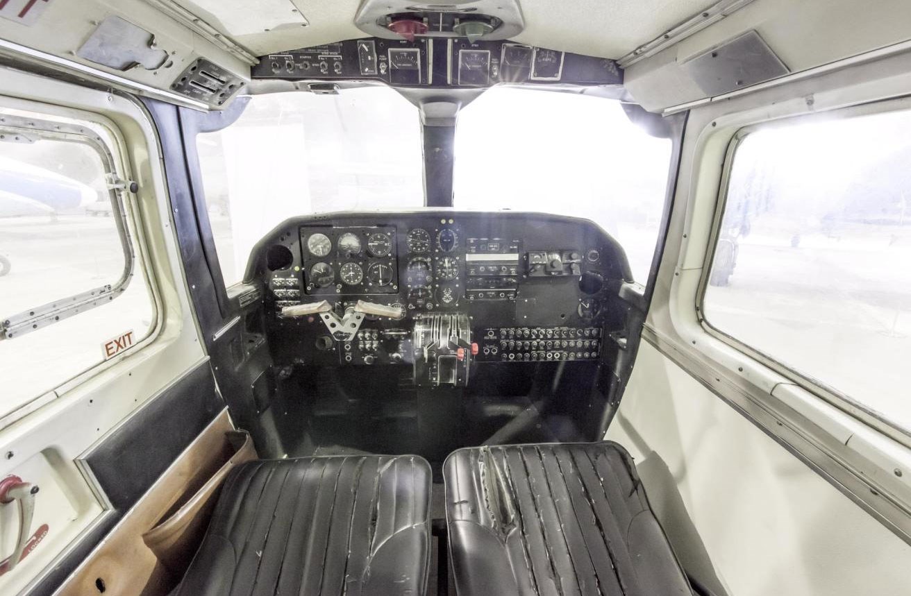 Inside the cockpit of a Britten-Norman Islander aircraft. There are padded black seats and the dashboard has several dials and buttons.