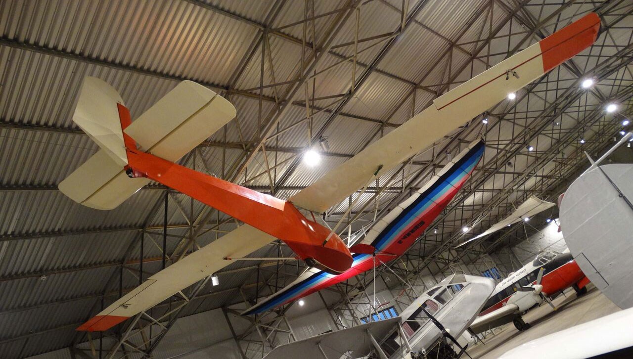 a red and white Rhonlerche glider hanging from the ceiling of a hangar, surrounded by other aeroplanes.