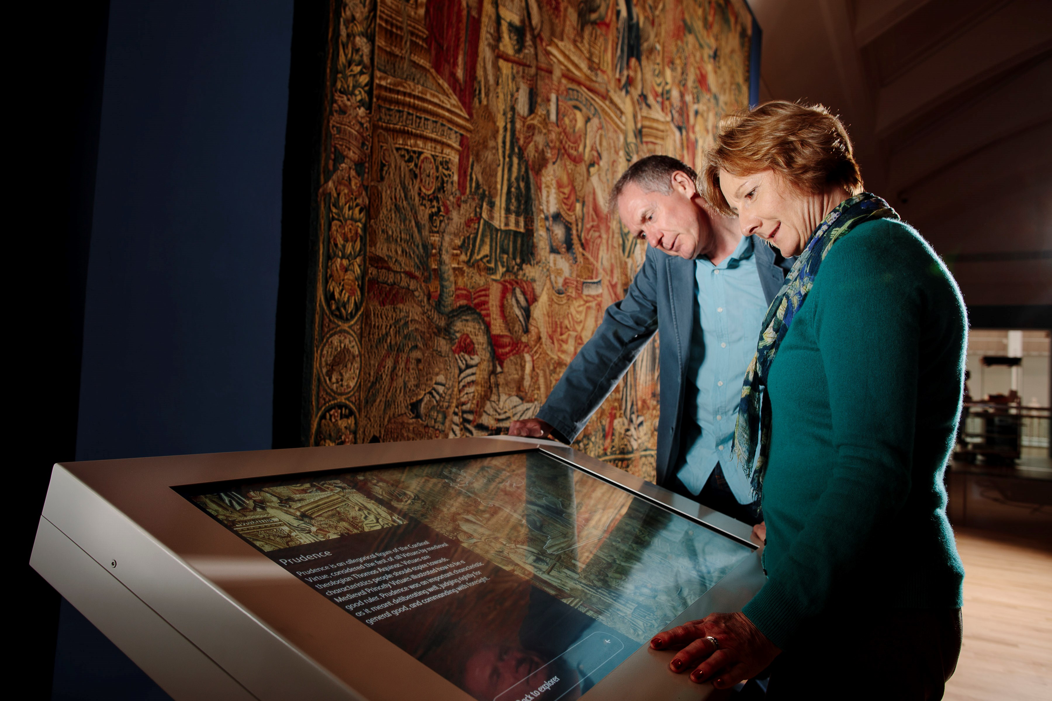 Two visitors looking at an interactive touch screen in front of a large tapestry.