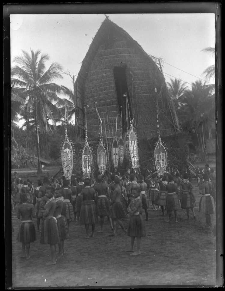A group of Elema people, standing in front of a thatched roof men's house, all wearing plant fibre skirts, neck ornaments, and nose ornaments, with several people holding hevehe (masks); Purari Delta, Papua New Guinea. © The Trustees of the British Museum (CC BY-NC-SA 4.0)