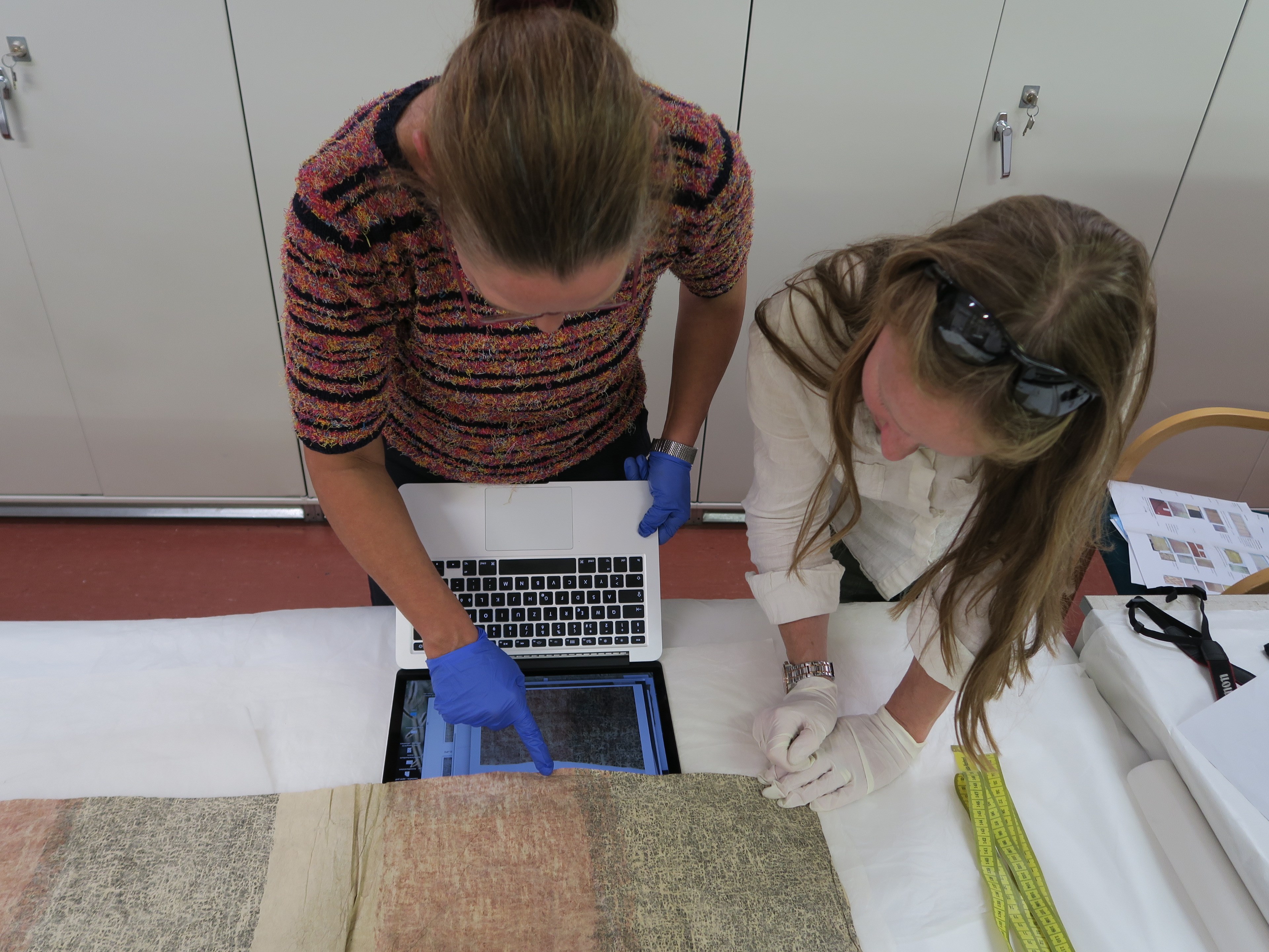 Antje Denner and Fiona Reilly leaning over a table comparing barkcloth 1881.2651 with a digital image of barkcloth A.UC.441.
