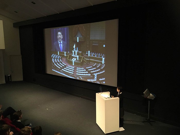 A student stand at a lectern on stage presenting slides on a screen behind and to her right.