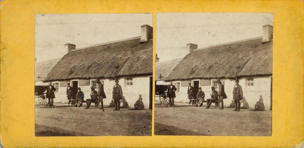 Yellow stereocard with two identical images of people standing in front of a single-storey thatched house.