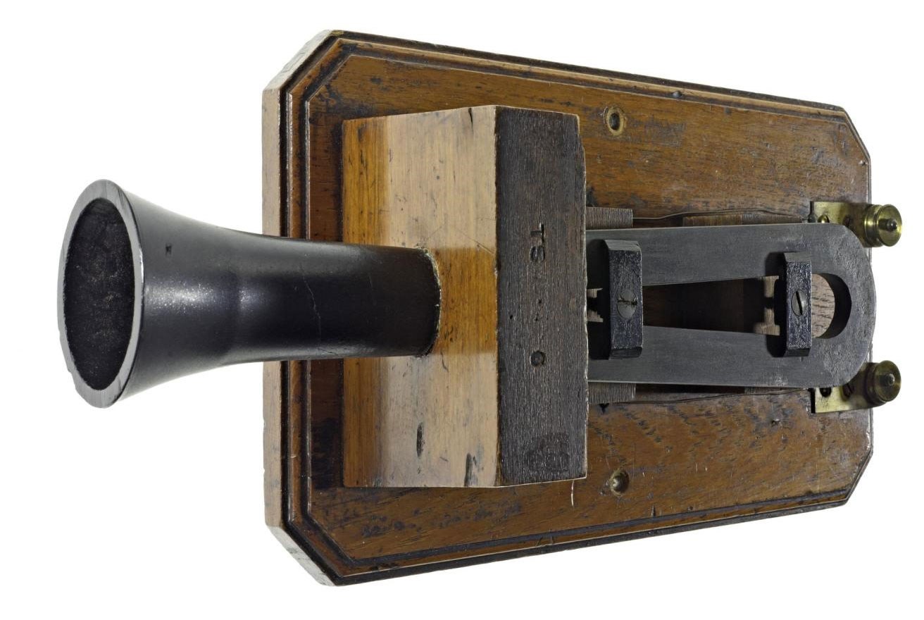 Box telephone on a white background. A wooden base with a black metal device and megaphone-like appendage sticking out.