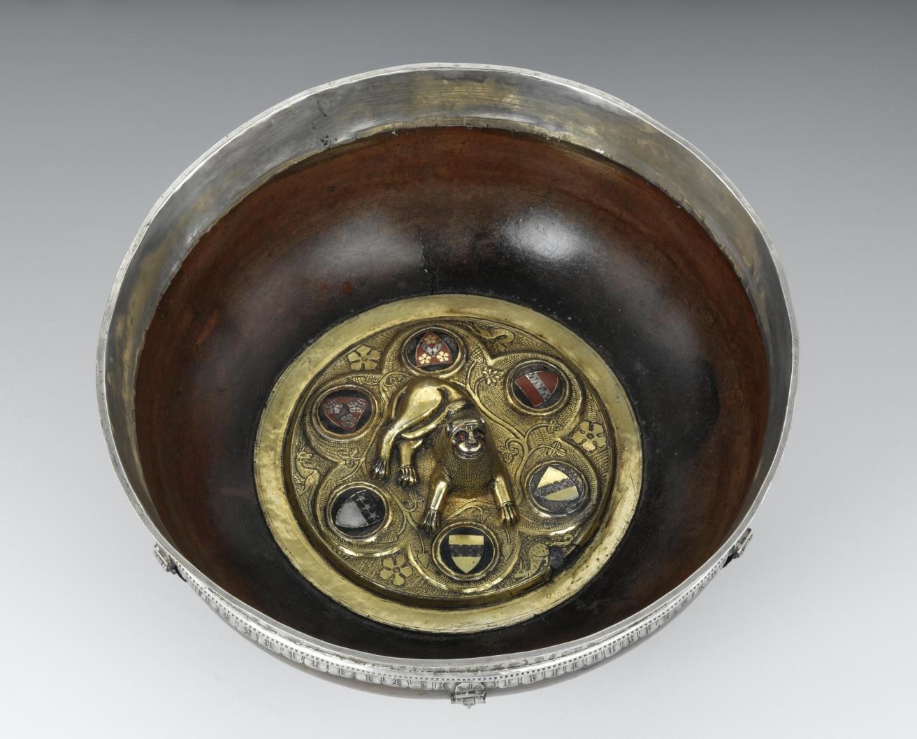 Inside of a wide metal-rimmed bowl or mazier. A golden disc at the base displays 6 coats of arms and a lion sculpture.