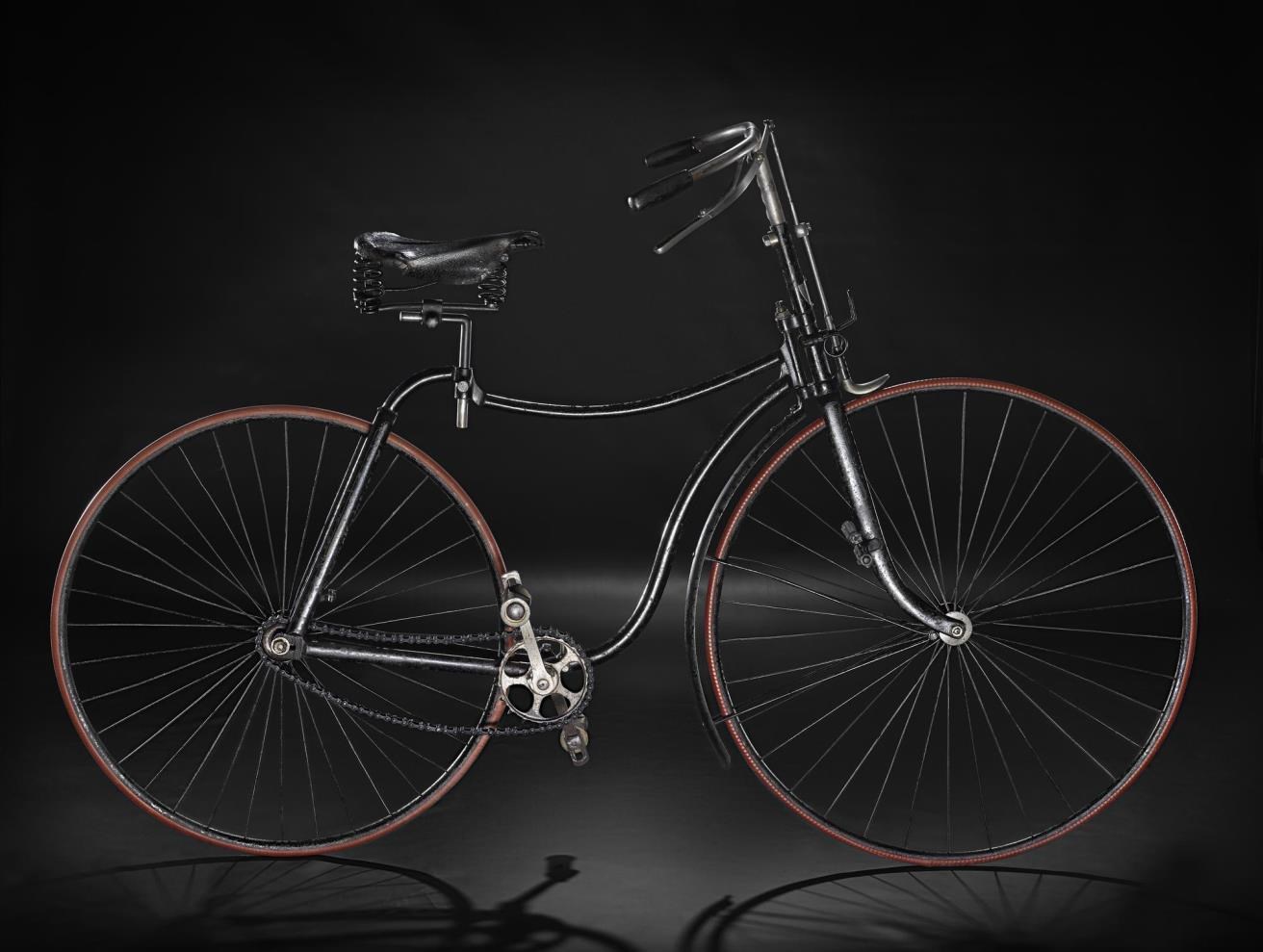 A vintage black bicycle with a light brown rim around the wheels, viewed from the side and standing upright against a black background.
