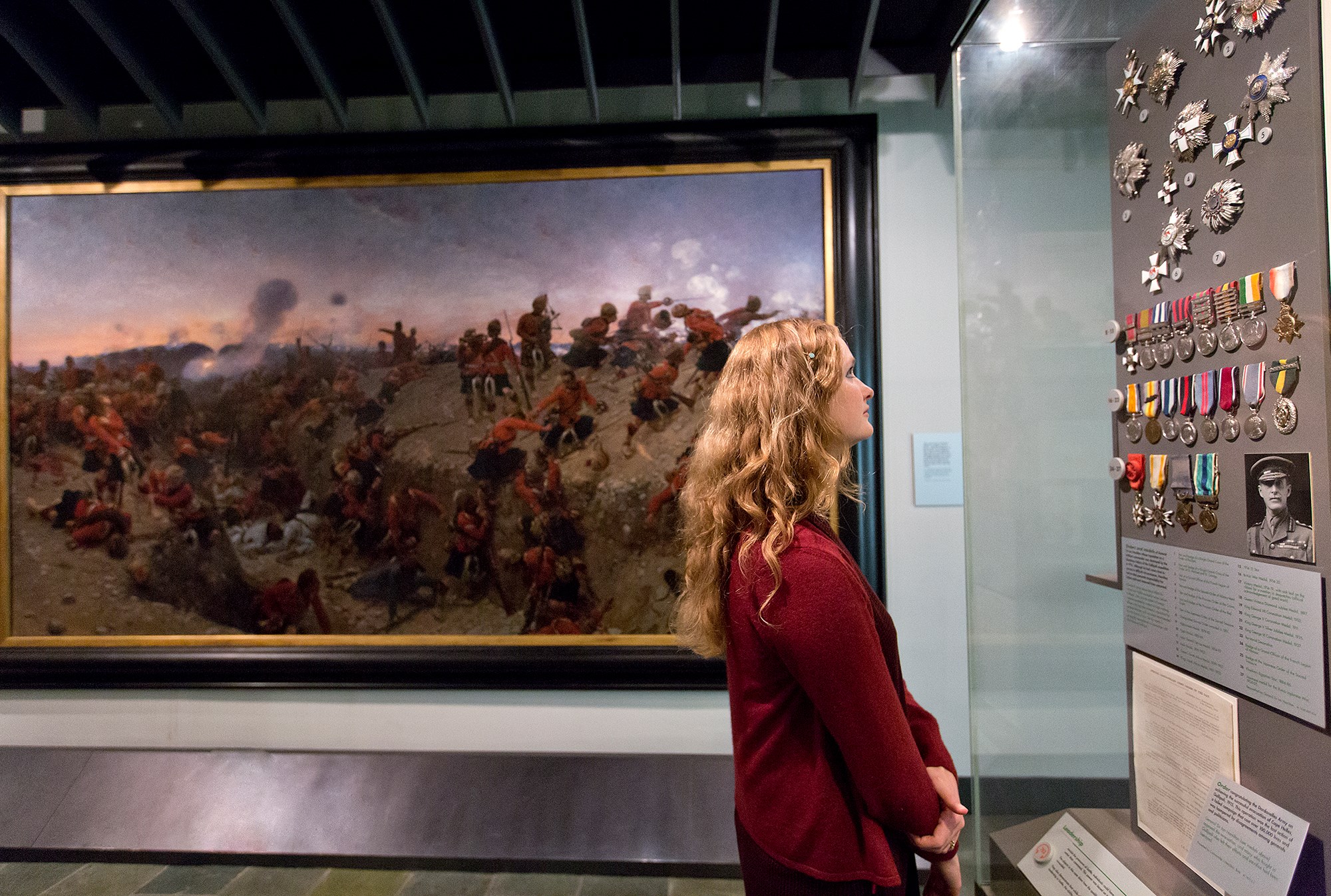 A visitor standing in front of a large painting of a battlefield looks at a display of medals hanging on colourful ribbons in a glass case.