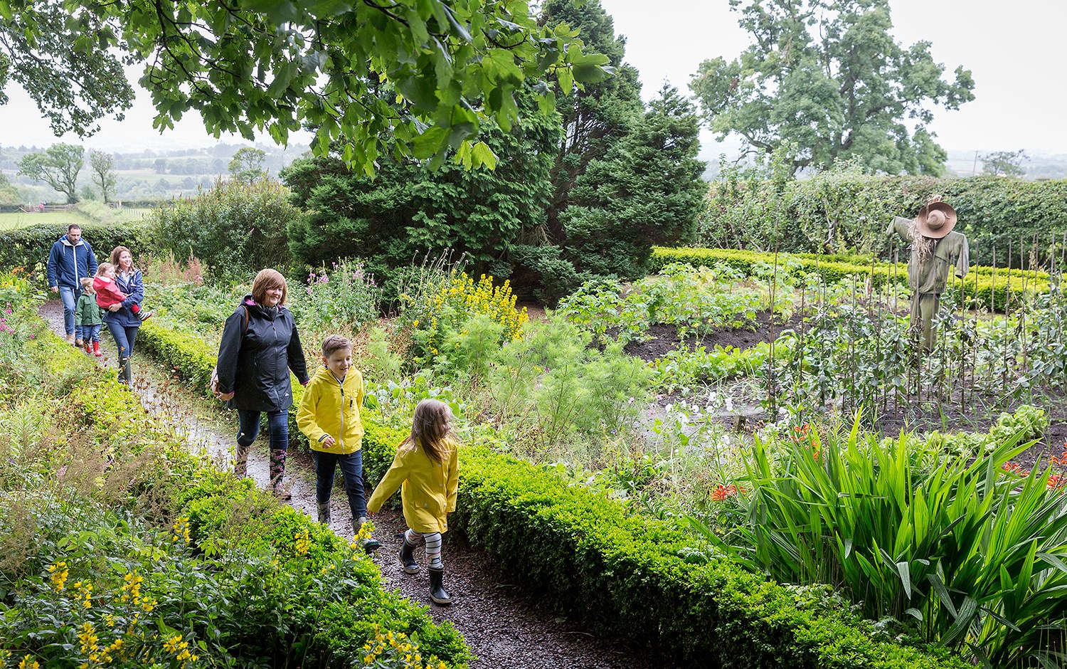 Two families walking along a trail in a garden with a scarecrow in the background. 