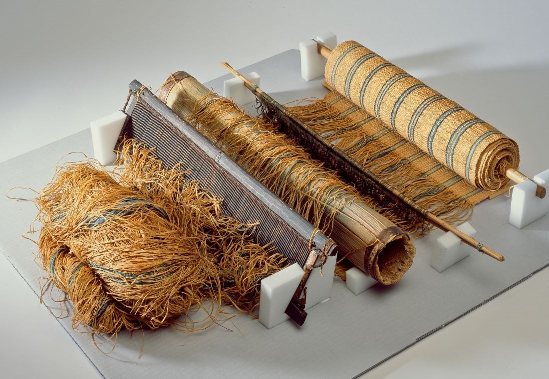 A small loom propped dup by several white blocks. Rough barkcloth strands on the left pass over a roller and comb-like barrier to emerge finely woven on the right.