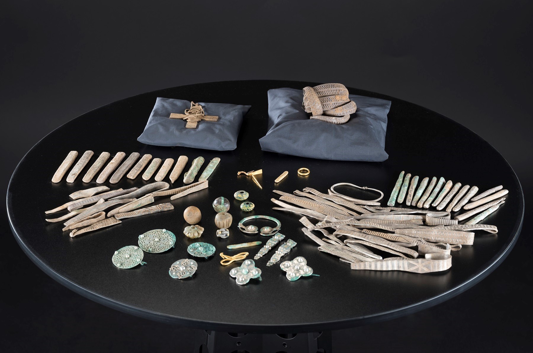 The contents of the hoard are laid out on a black table. Pillows prop up the cross and arm-ring bundle with the rest organised by object type.
