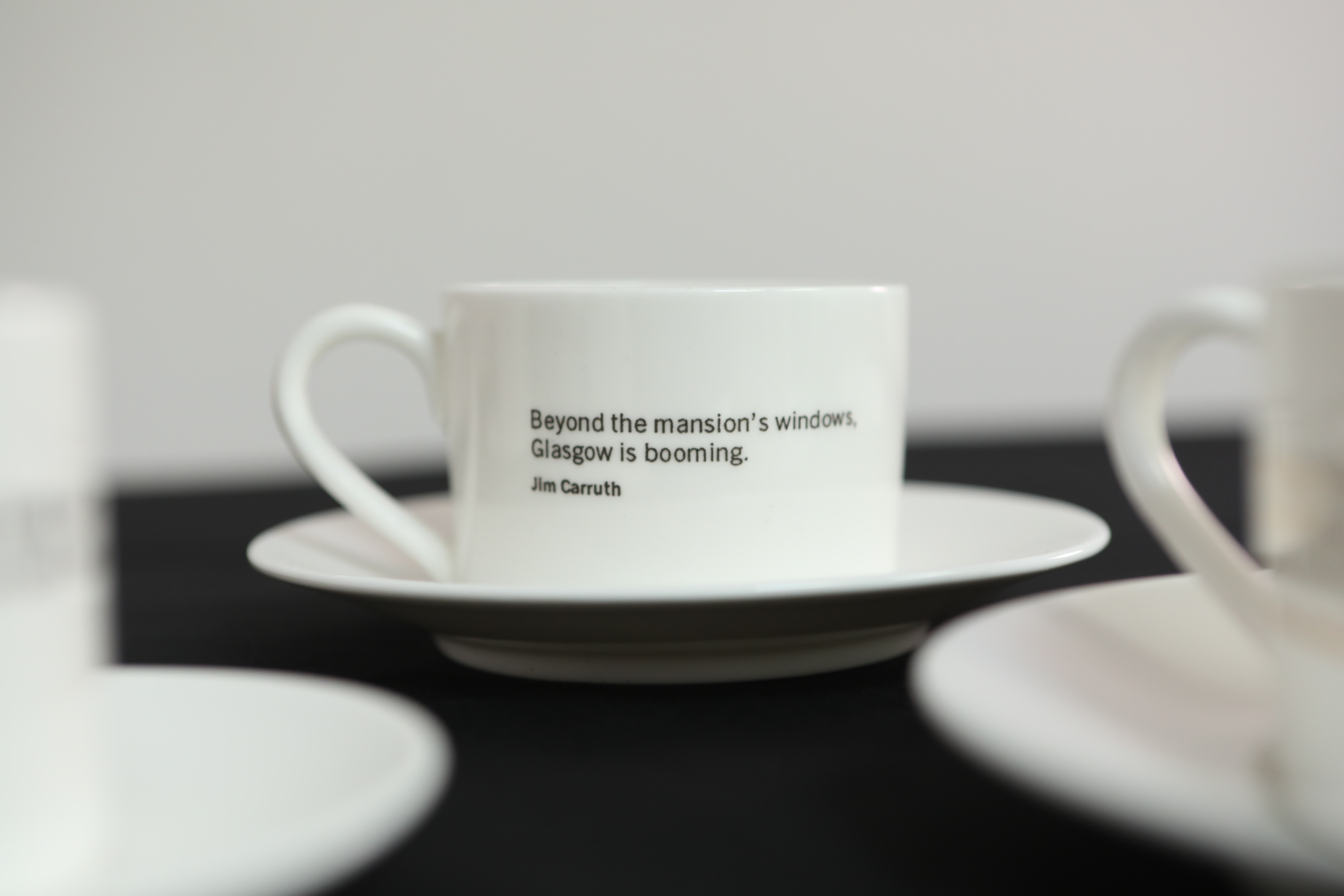 White cup on a saucer with quote printed on the side - 'Beyond the mansion's windows, Glasgow is booming.' Jim Carruth