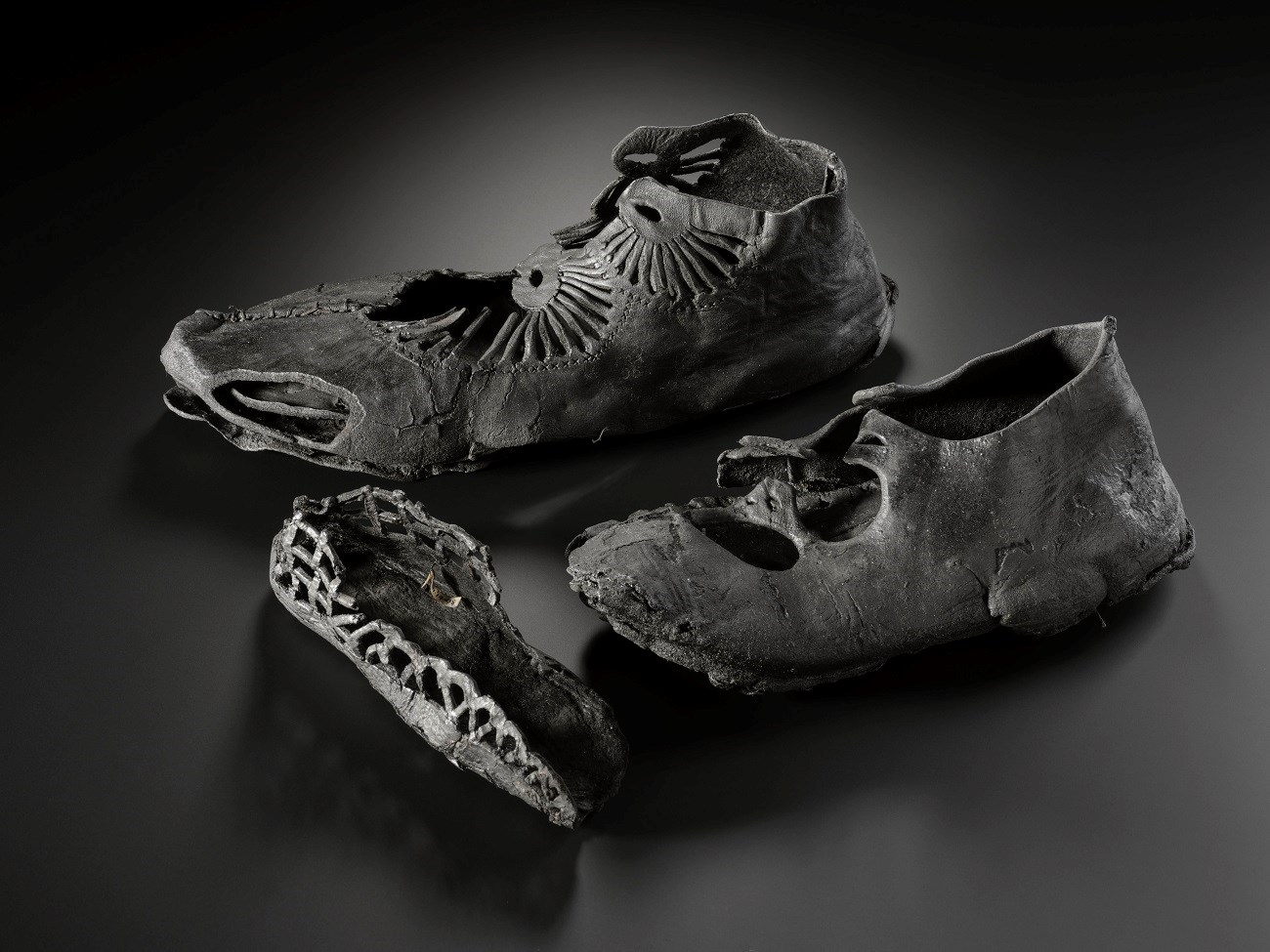 Three shoes on a black background. One is large with a radial pattern of straps, one is medium with less complex straps, and one is small with just the sole surviving.