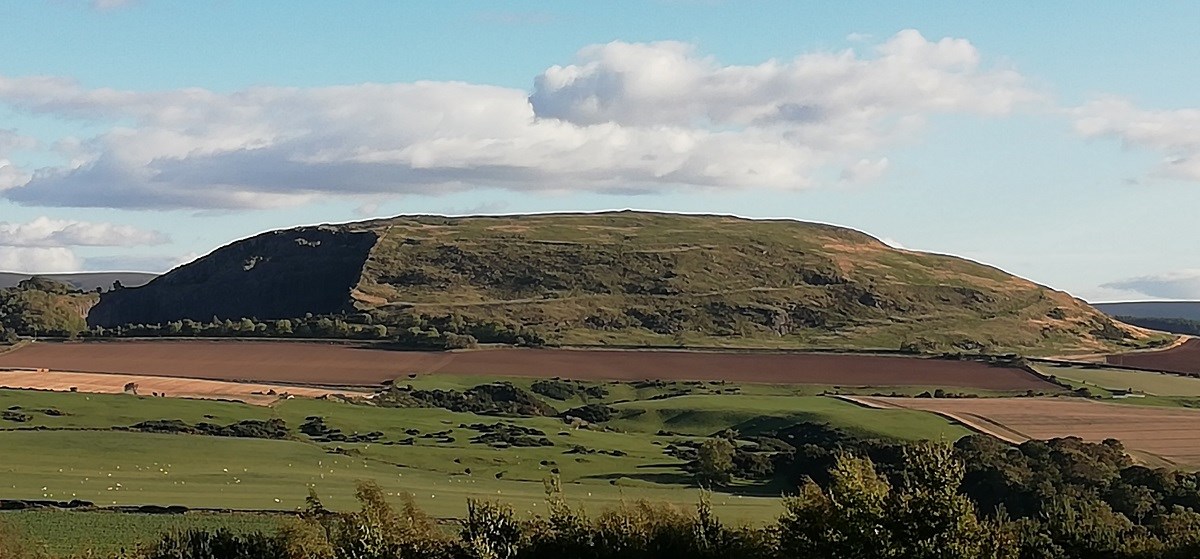 A whaleback-shaped hill rises up from fertile fields under a cloudy blue sky.