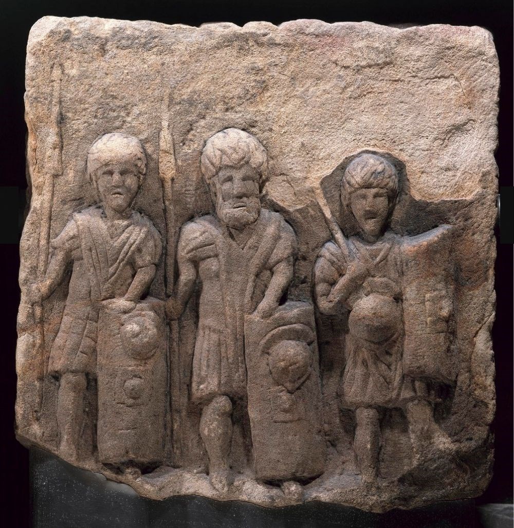 Stone block depicting three boldly carved figures of soldiers standing side by side and looking straight ahead, armed with spears and shields. They feel like friends.