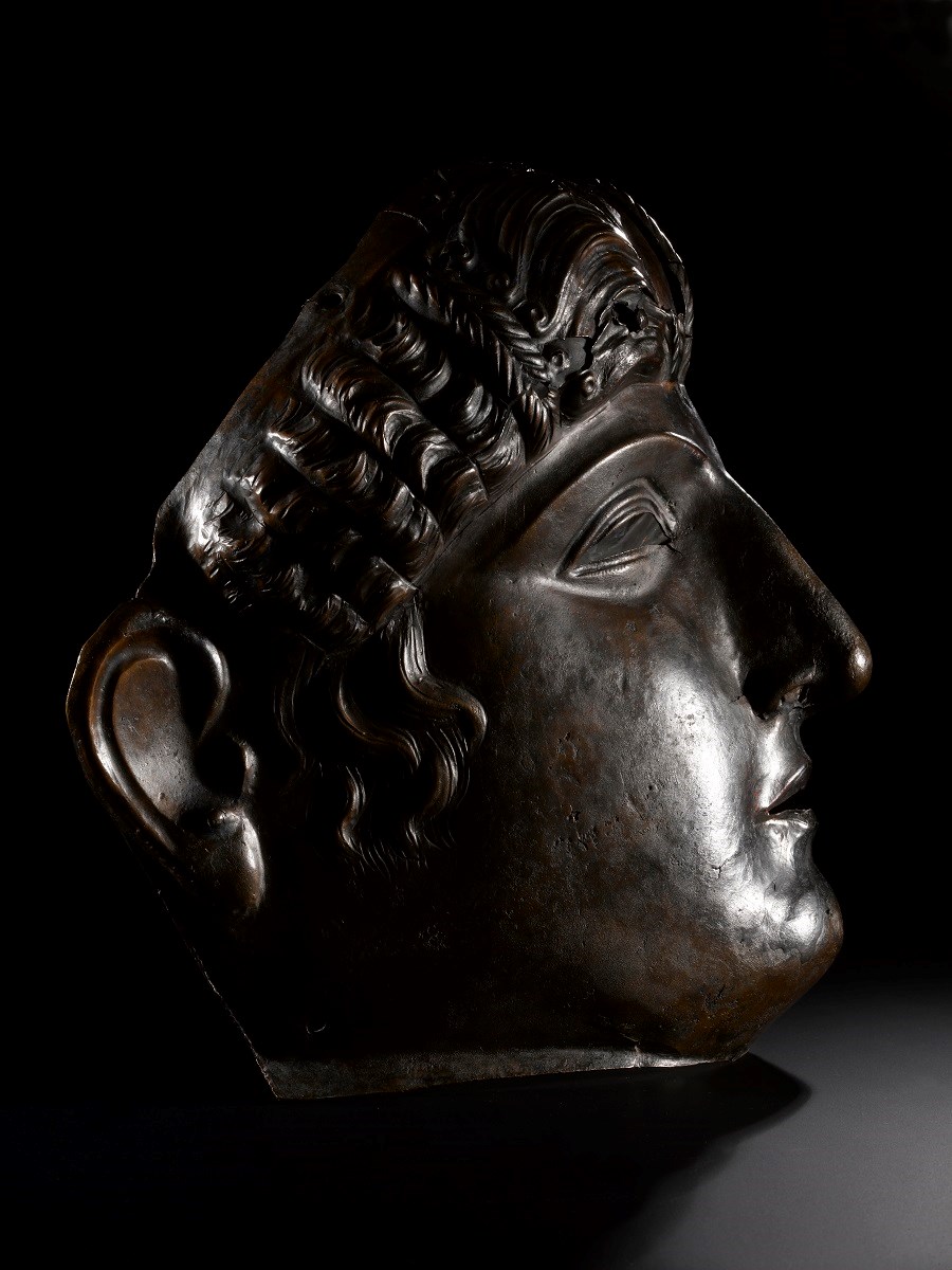 Profile view of bronze mask placed on a table. Light glints off its right cheek, and it bears a dignified expression.