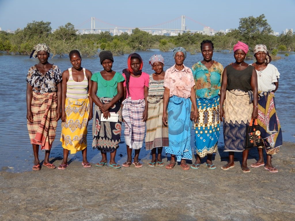 Group of nine women standing on the shore of a river on a sunny day. All facing forward, colourfully dressed. 