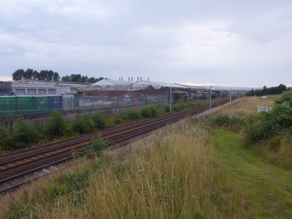 A field is abruptly halted by a train track, with industrial buildings on the other side.