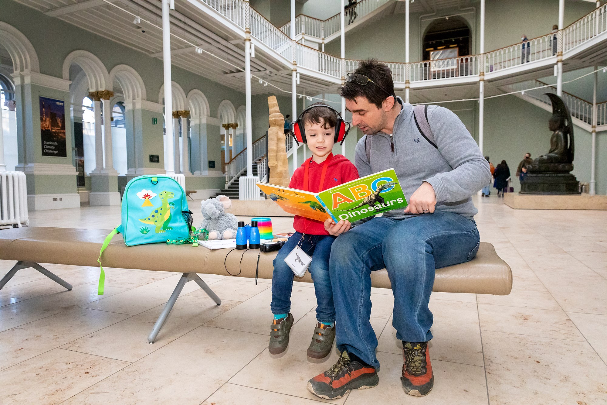A man and a boy sit on a bench in the grand gallery looking at a large dinosaur picture book. The boy is wearing ear protectors.