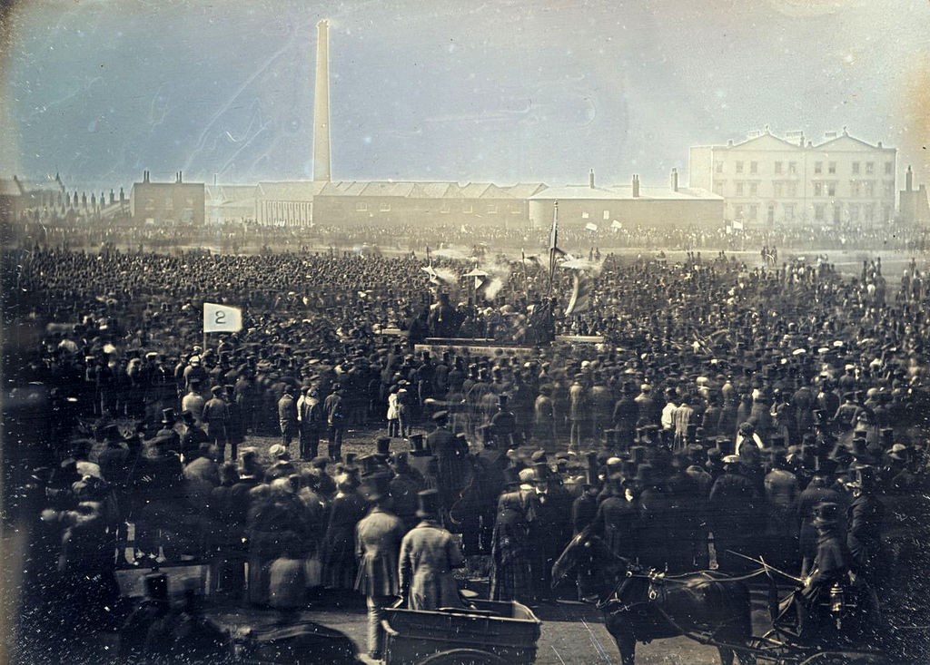 Vintage photograph damaged by light of a massive crown in top hats, suits and dresses gathered near a factory in London.