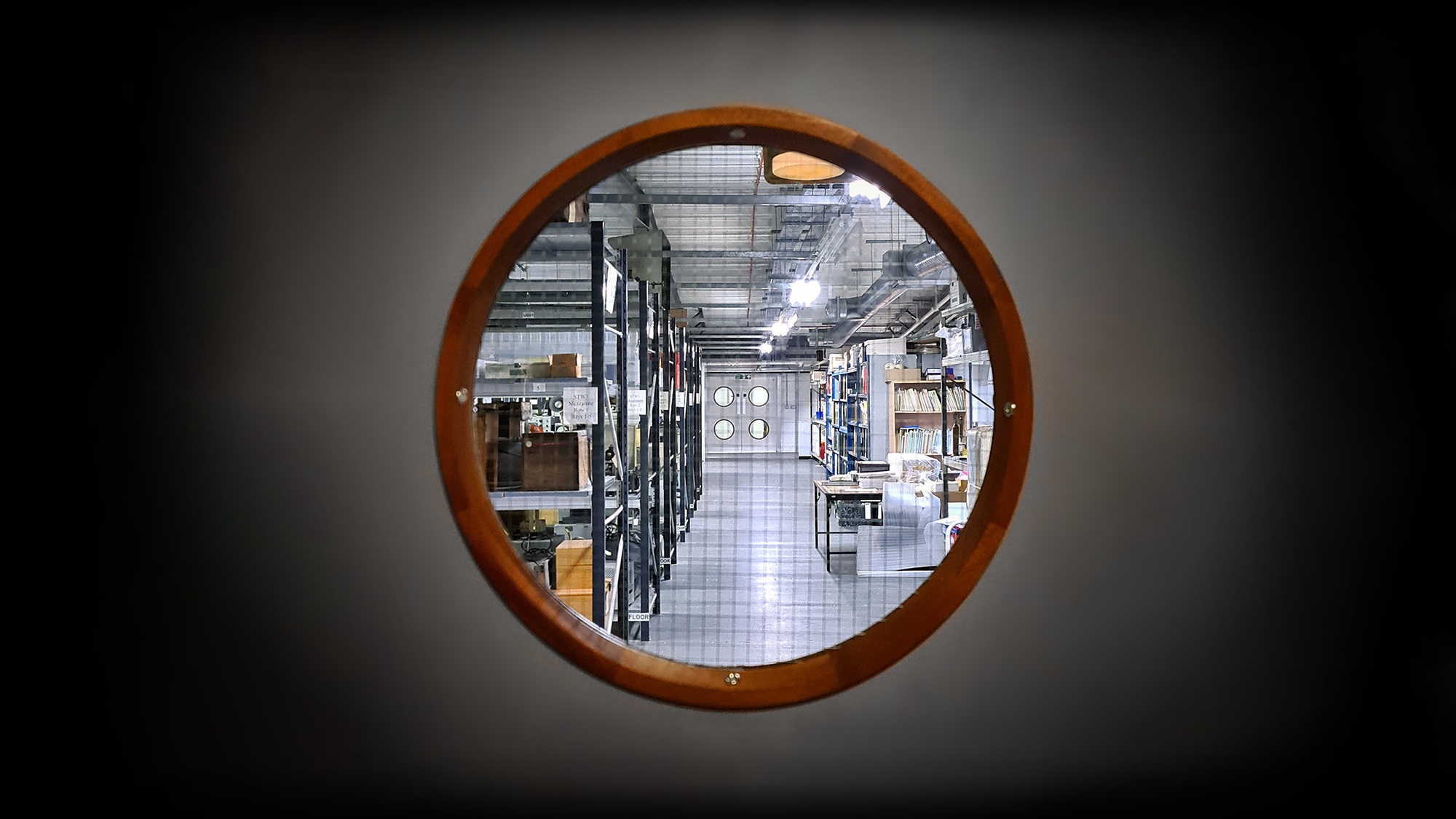 A small circular window, like those you would see in the hull of a ship, gives a glimpse into a museum storage area filled with racks containing boxes and folders.