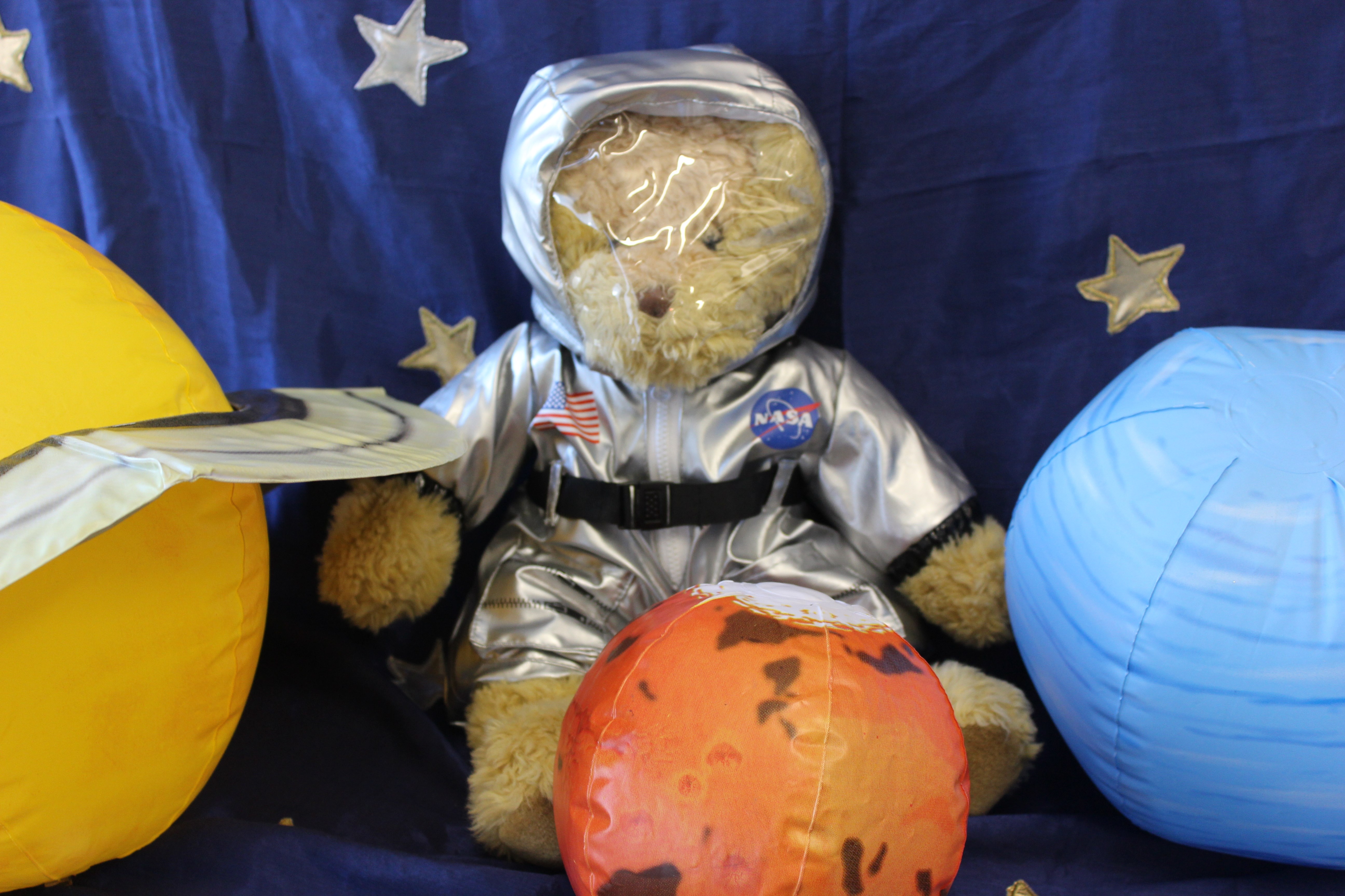 A teddy is dressed in a silver space suit. It is placed against a start background and surrounded by inflatable planets. 