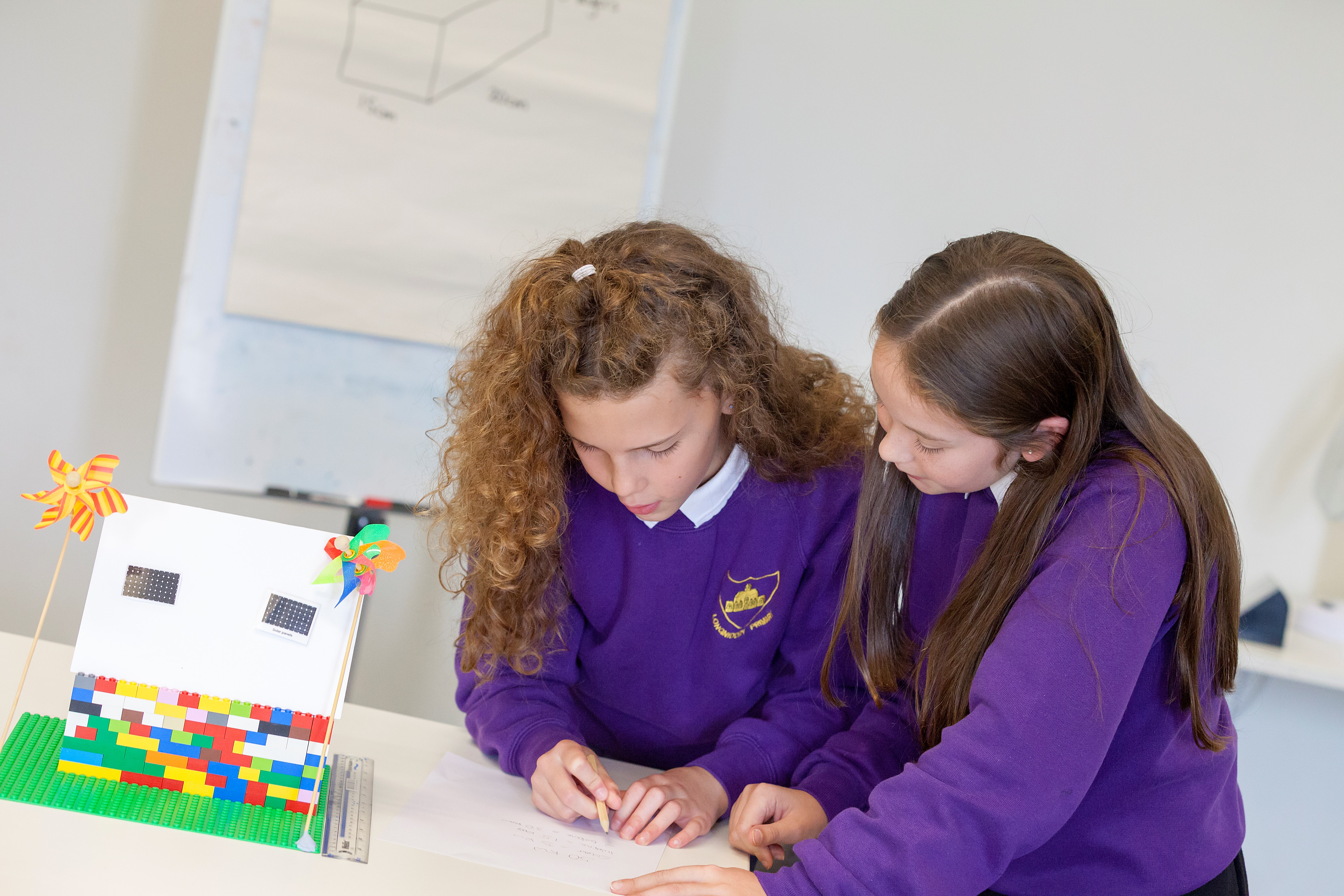 two pupils do some calculations on paper next to a small lego house, which has mini solar panels on the roof and windmills outside. 