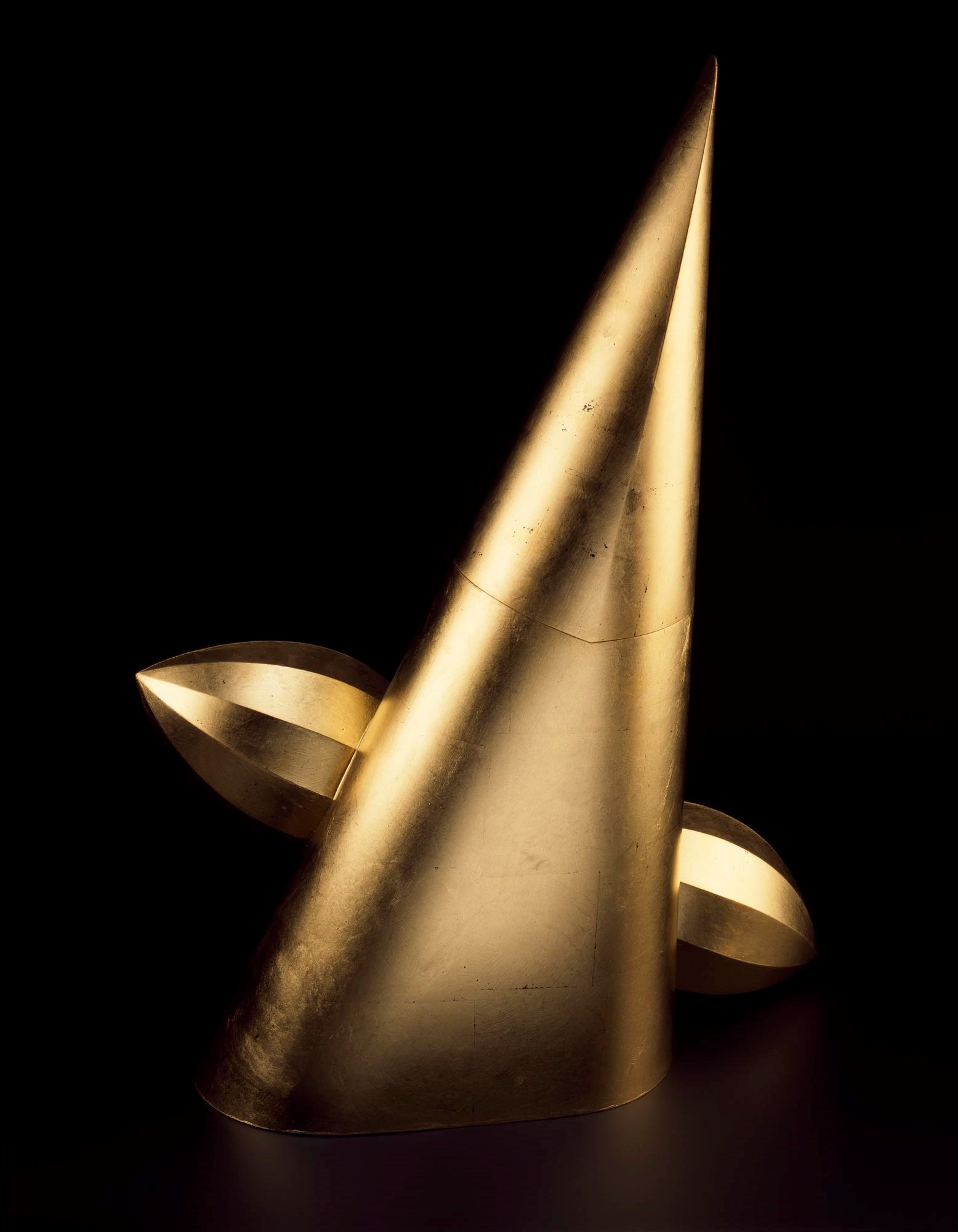 Metal sculpture, golden in colour. A tall triangle, becoming conical at the bottom and pointy on top, is pierced by a bullet-like pointed and flat-sided golden tube.