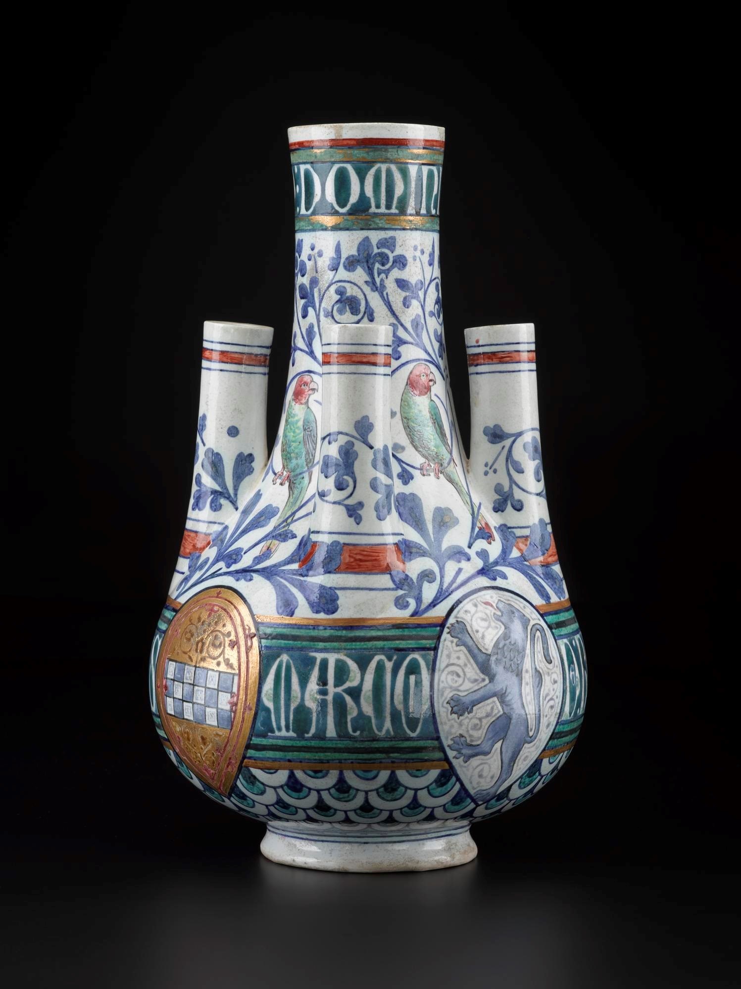 Flower vase with one large, central cylinder and three shorter, vertical spout. Decorated with coats of arms, blue leaves, Latin letter and green parrots.