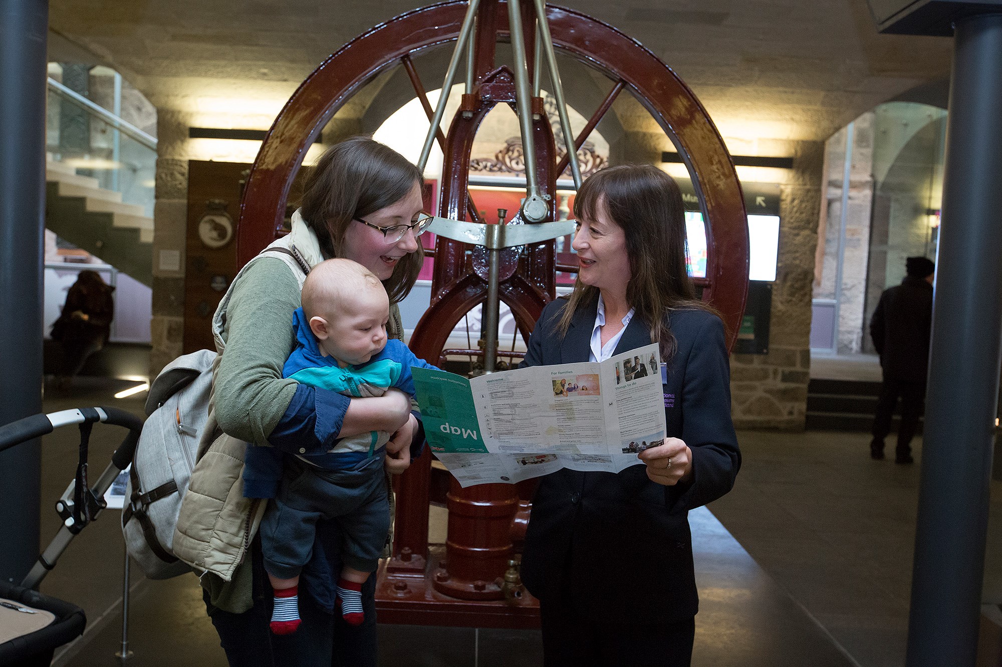 A woman holding a baby looks at a map held by a member of the visitor experience staff.