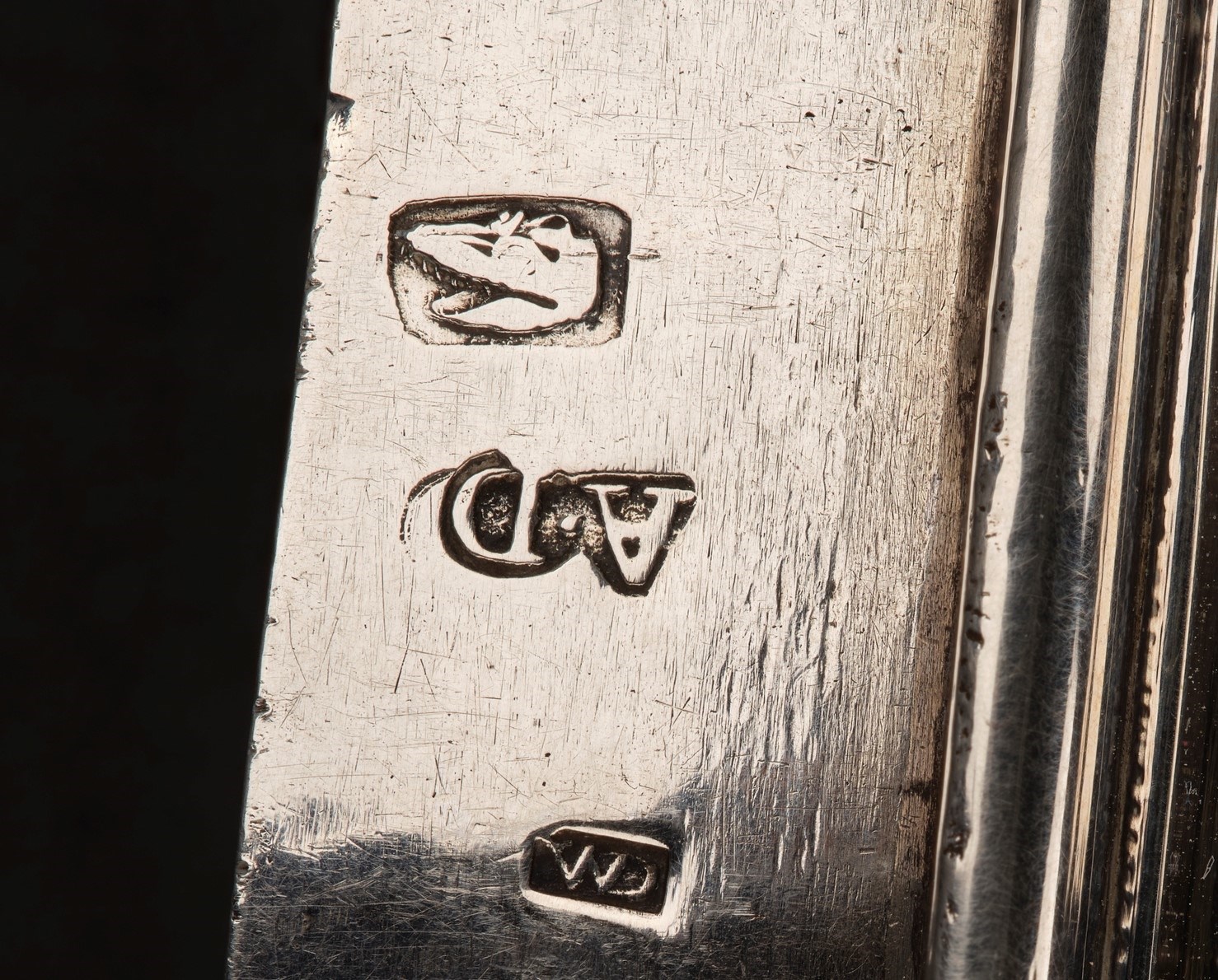 Closeup of a stamped engraving on the edge of a silver vessel. There are three stamps: on top, an alligator's head within a rectangle. In middle, the letters 'A' 'D' upside down. On bottom, the letters 'C' 'M' updside down.