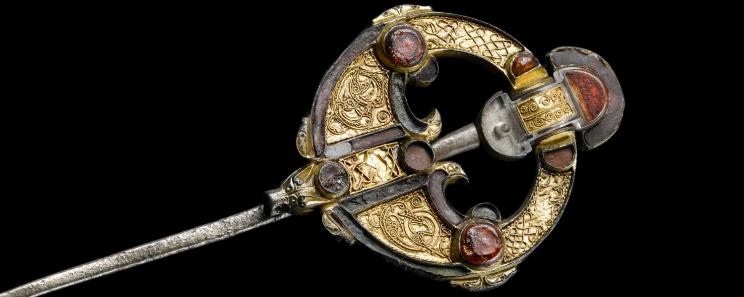 Gold-coloured brooch and pin, intricately decorated with raised Pictish-style patterns and red gemstones.