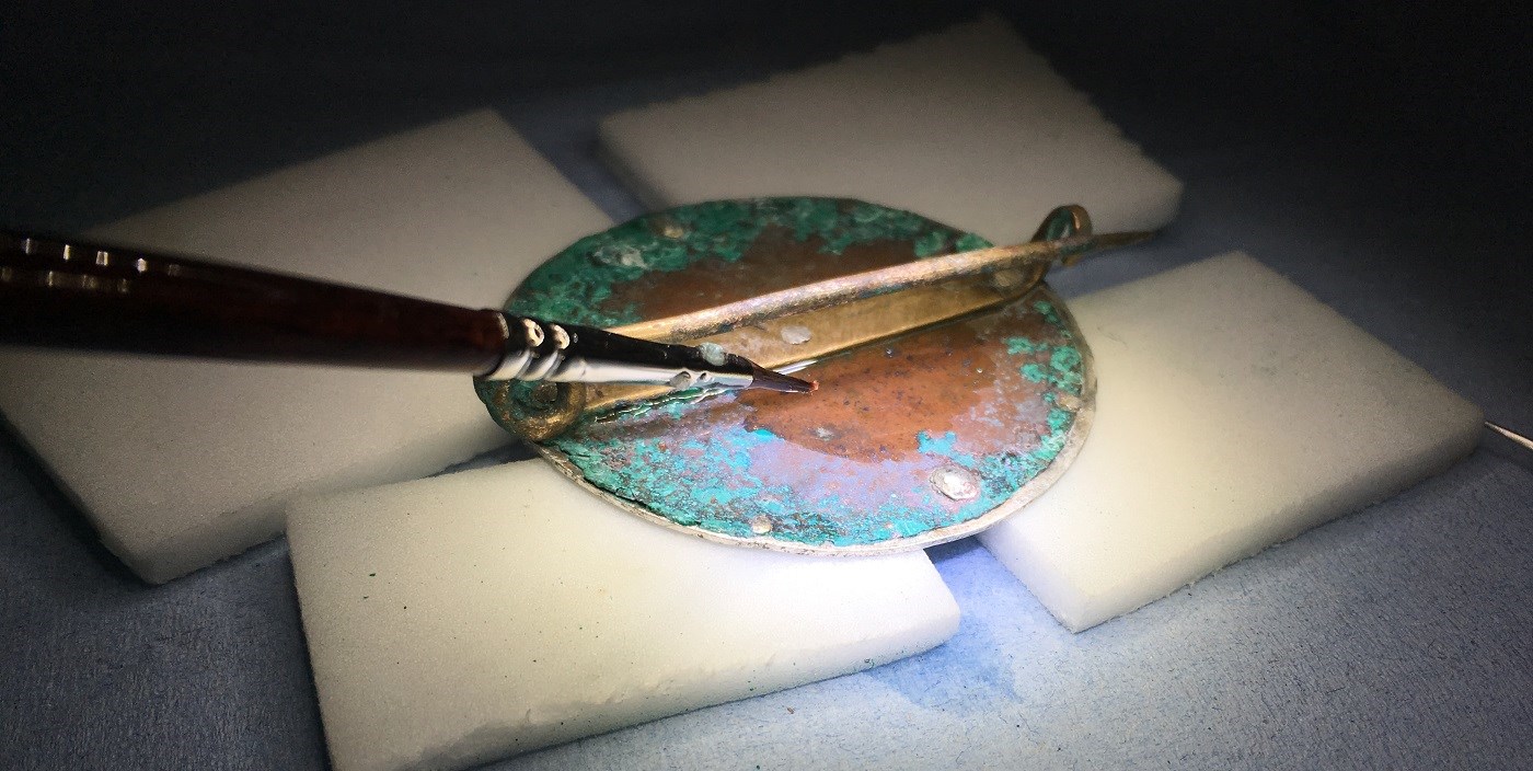 The circular brooch rests atop four rectangular pads under a bright light. A metal tool is being used to delicately remove corrosion from its copper and green surface.
