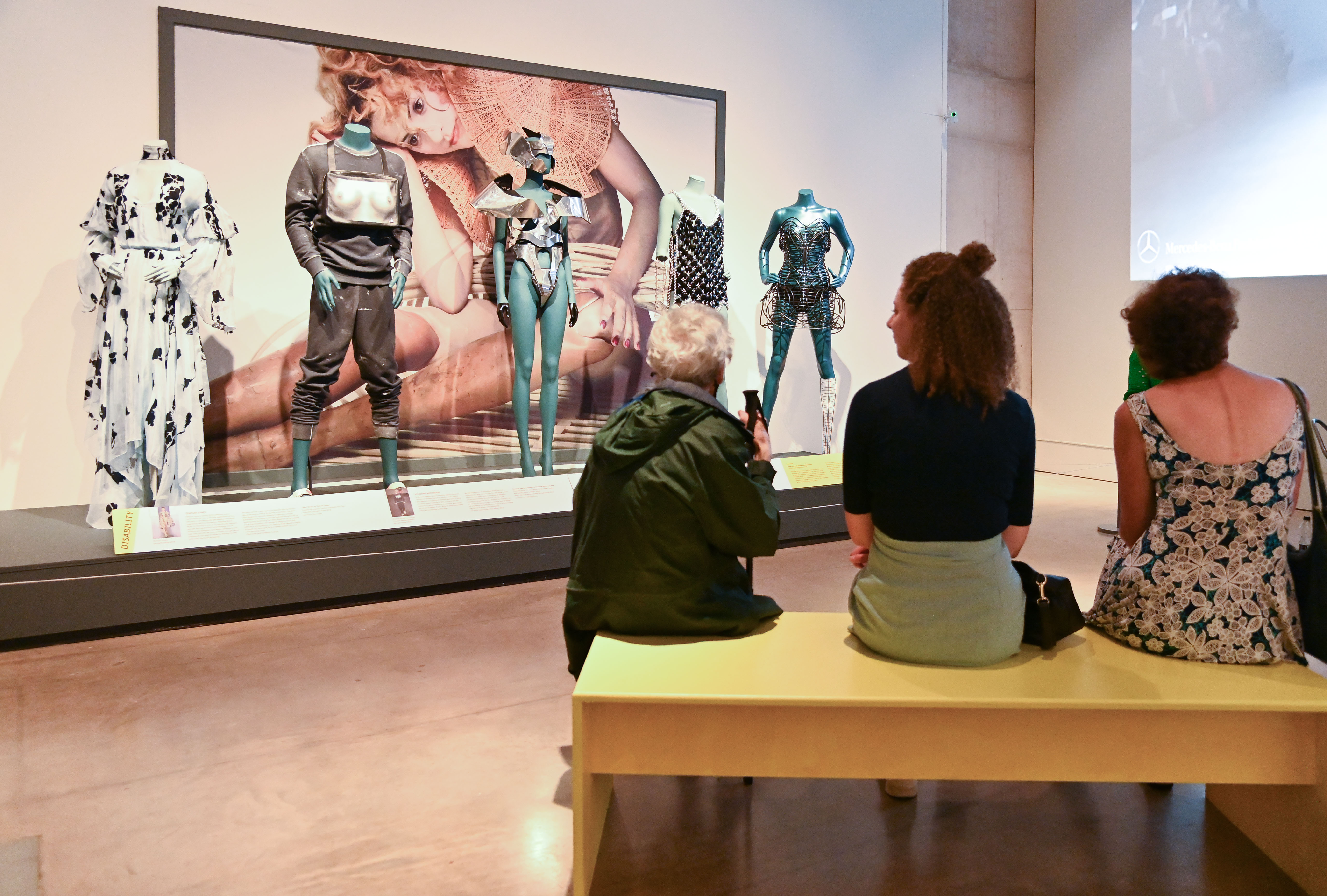 Three visitors sit on bench and look at five styled mannequins within the exhibition.