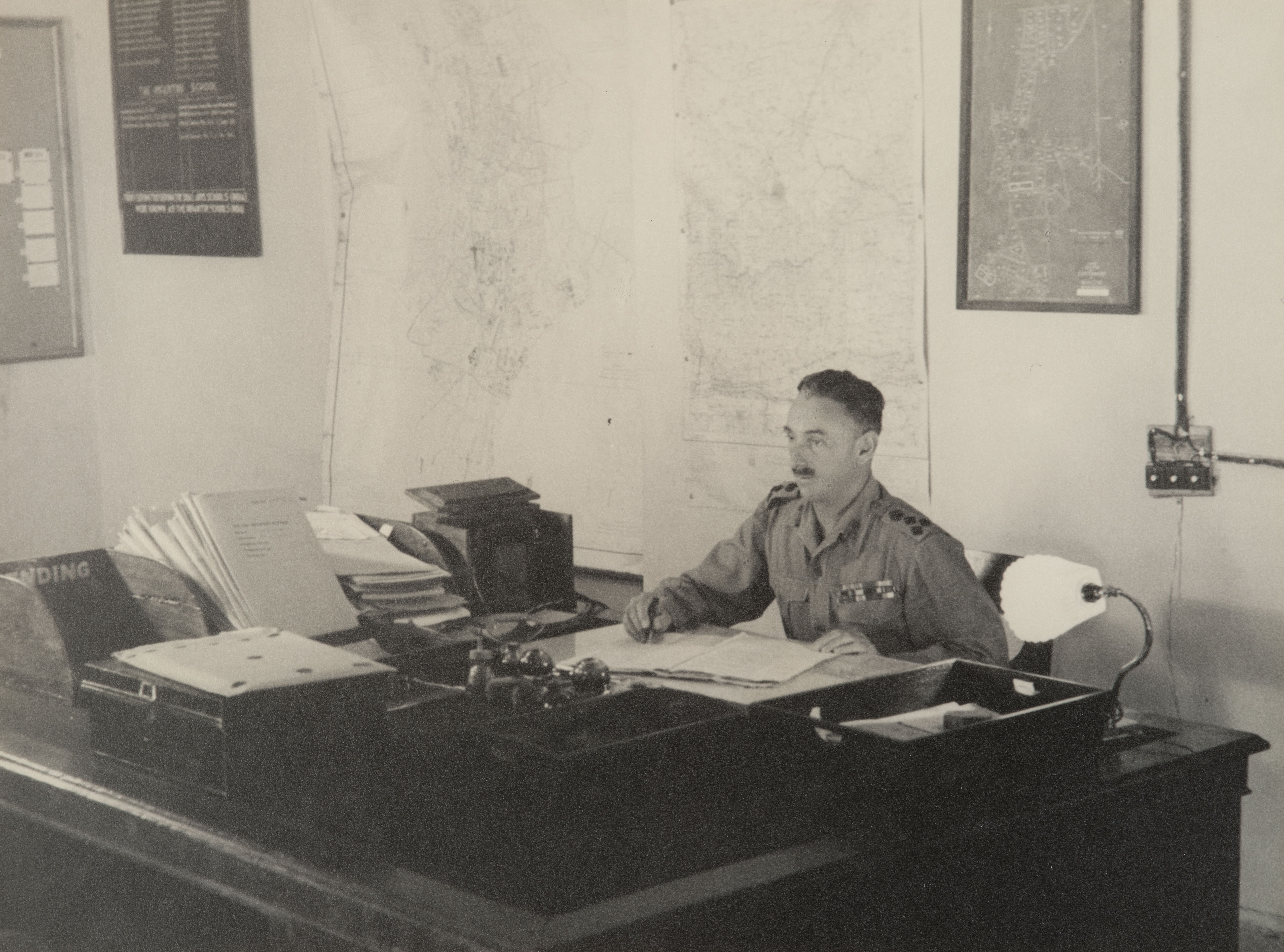 Brigadier Norman Macdonald sits at a desk surrounded by papers.