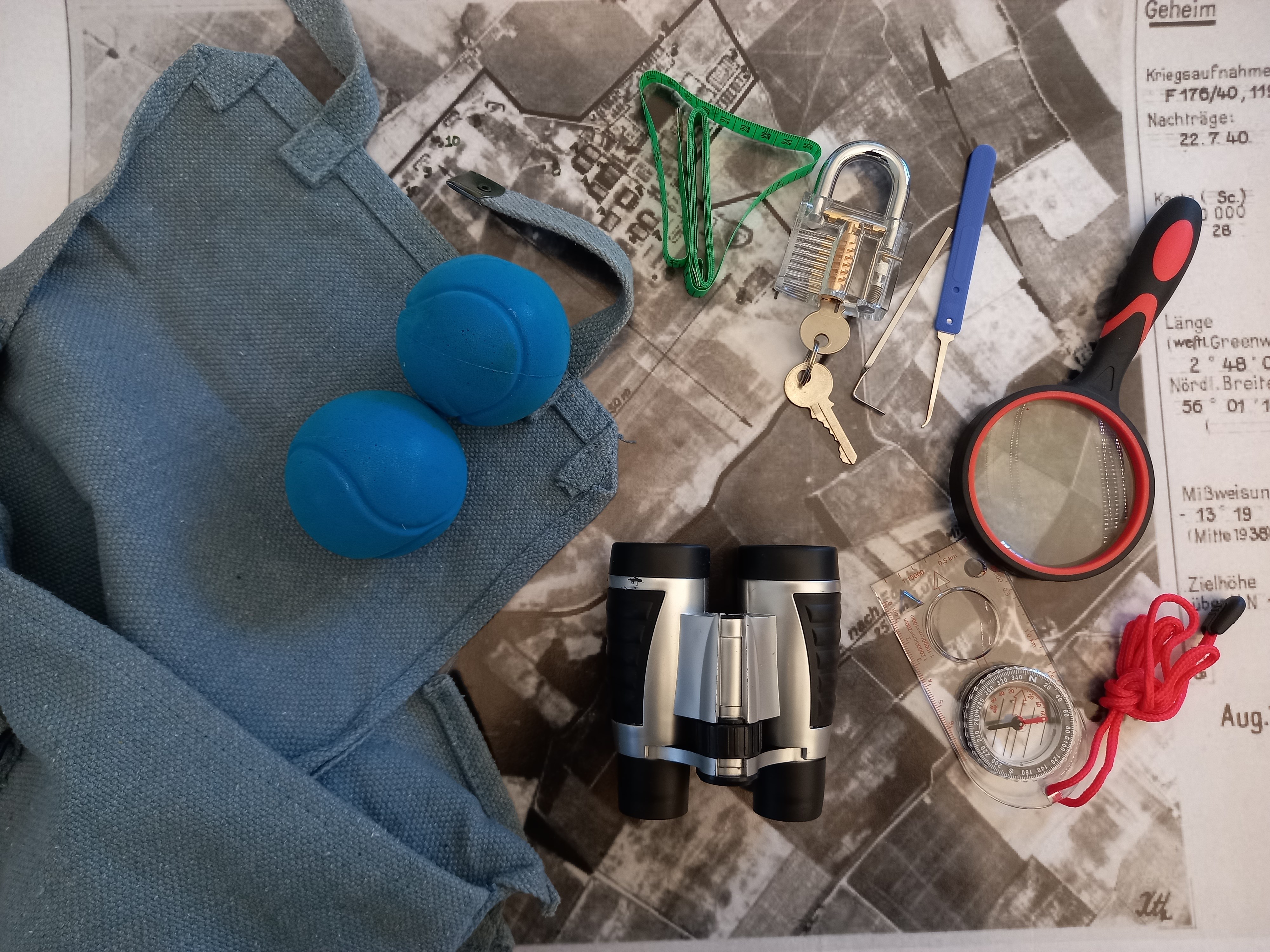 Blue tennis balls, binoculars, magnifying glass, compass and lock and key lie beside blue satchel bag.