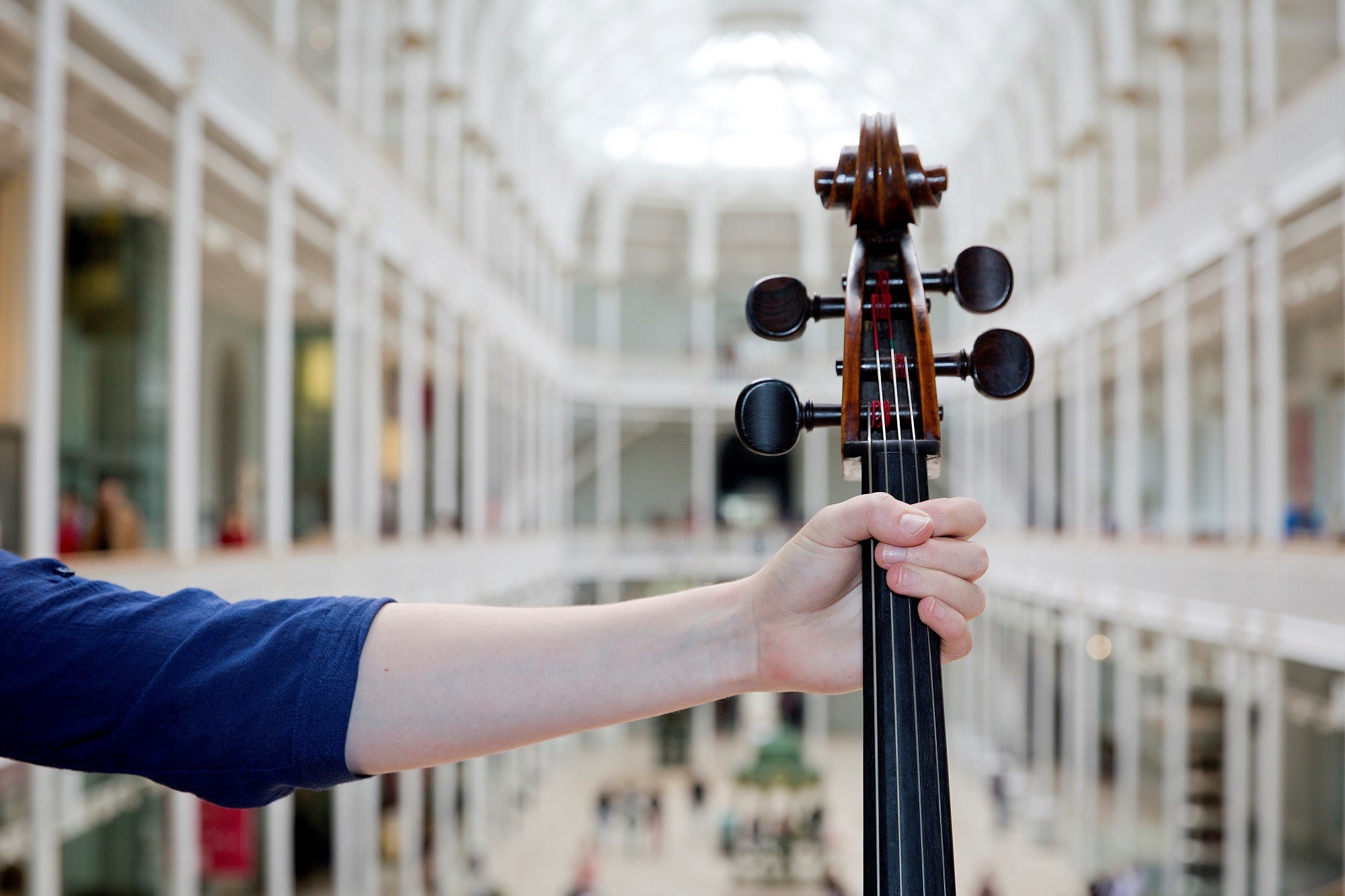 A hand holds the neck of a violin in front of the grand gallery at the National Museum of Scotland.