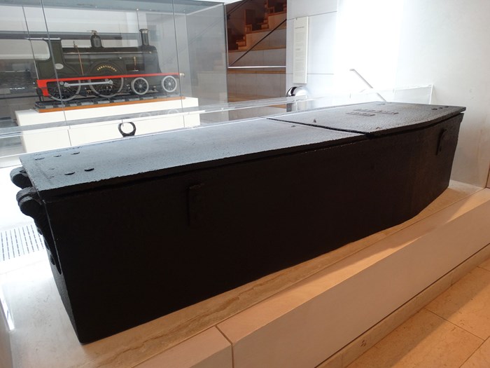 Large black metal crate shaped like a coffin. Set in a museum display area.