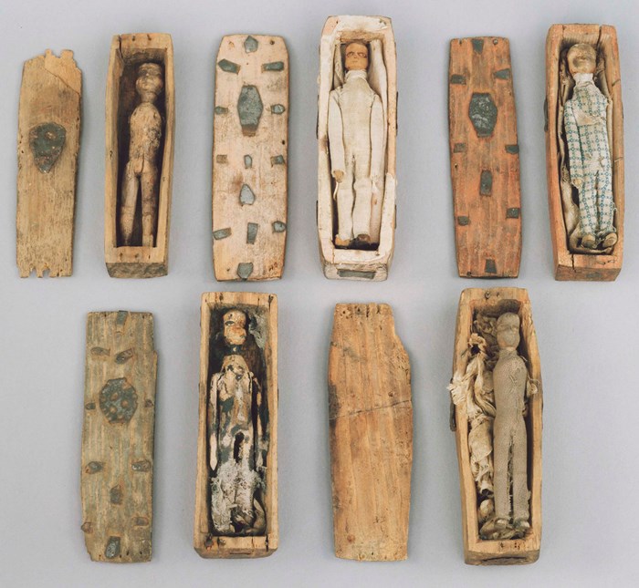 Top-down view of five of the Arthur's Seat coffins. Light brown coffins with skinny wooden dolls inside them, lids open, on a white table.