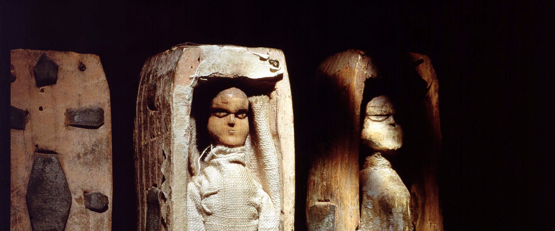 Two coffins with dolls inside, upright against a black background with heavy shadows cast over the coffins to create a spooky atmosphere.
