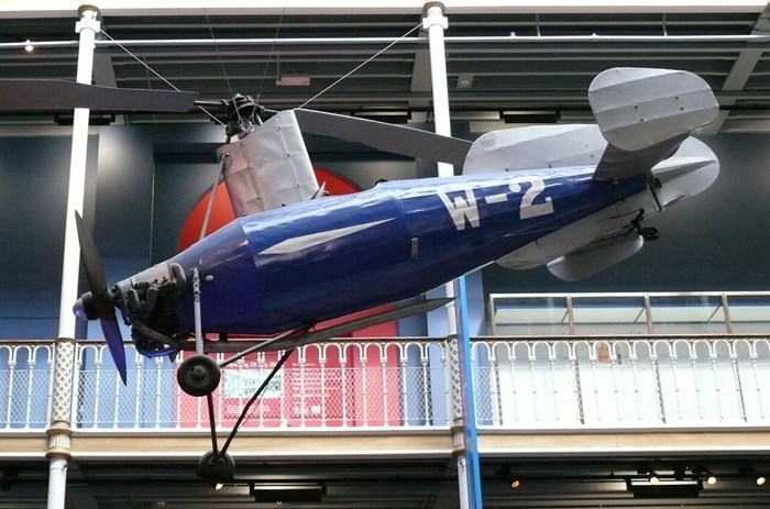 The sideview of a blue and white Weir W-2 autogyro aircraft suspended from the ceiling of a museum gallery.