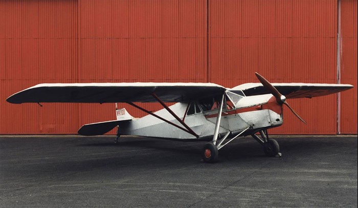 A white de Havilland Puss Moth aircraft parked in front of a red hangar.