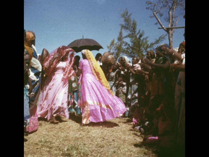 Slide of women dancing as others clap, an umbrella is held above the dancers and they are wearing brightly coloured clothes: Jijiga, Ethiopia, 1960s, photographed by Jean Jenkins.